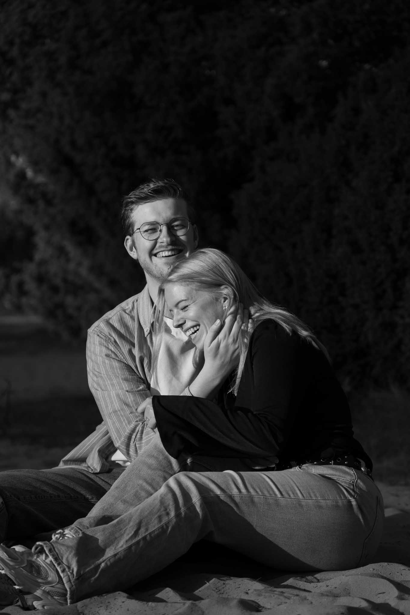 Een koppel zit in het zand waarbij de man lachend naar de camera kijkt en de vrouw lachend tegen de man aan leunt