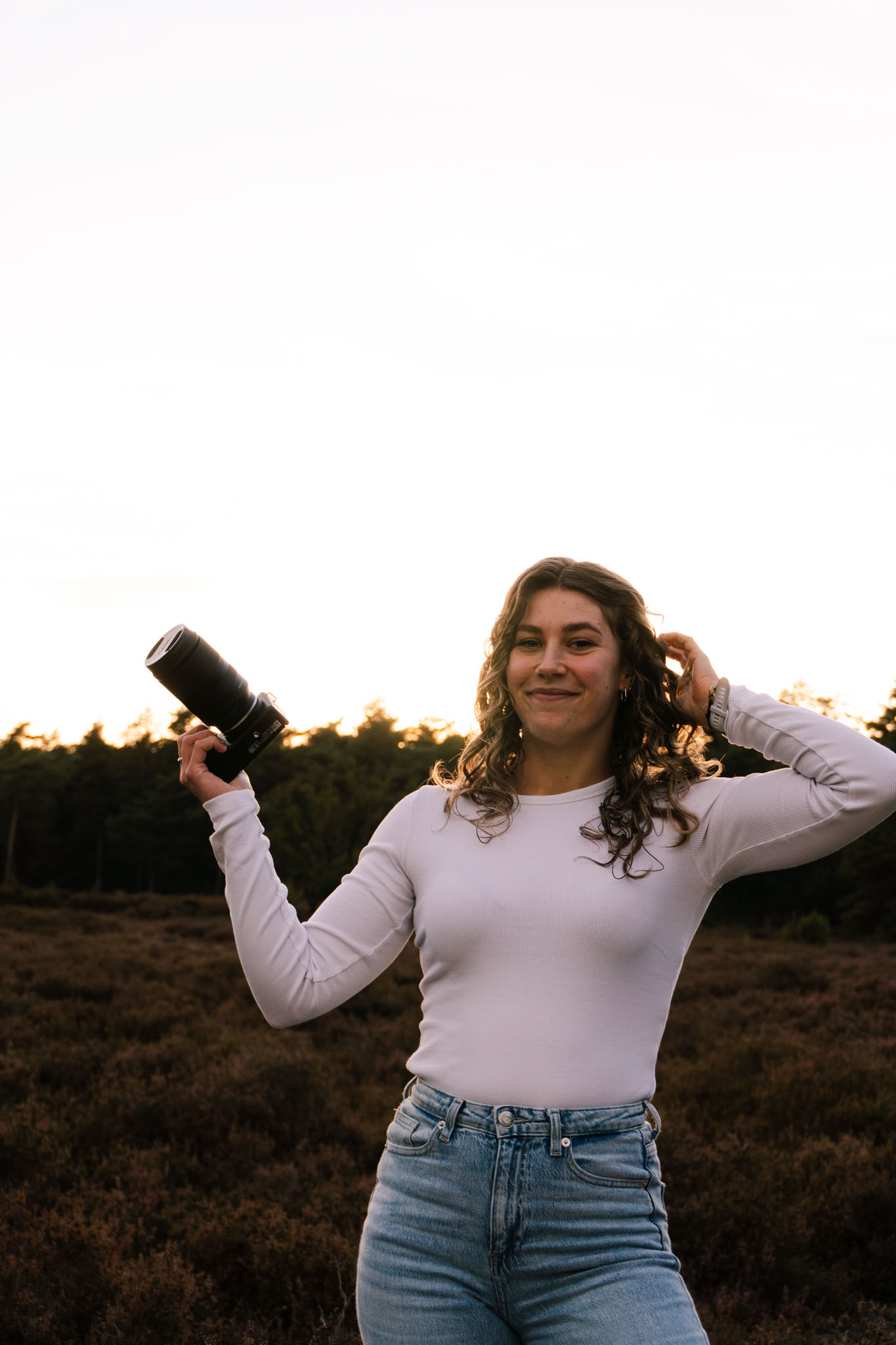 fotograaf lachend richting de camera. ze gaat met haar ene hand door haar haar, met haar andere hand houd ze een camera omhoog. ze staat tussen de mooie heide