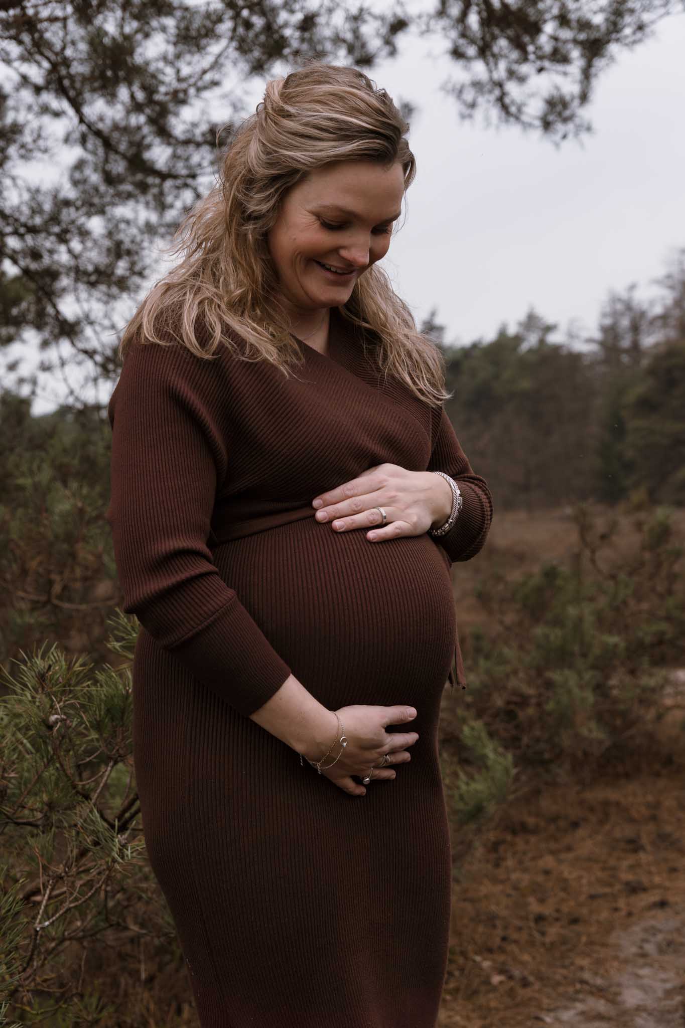 Zwangere vrouw kijkt lachen naar haar bolle buik.ze draagt een strakke bruine jurk, waarin de zwangere buik goed te zien in.