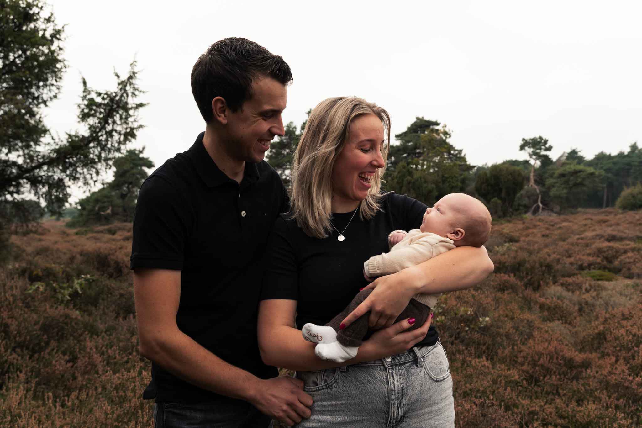 liggende foto van een jong gezin met pas geboren baby. ouders kijken lachend naar de baby die bij moeder in de armen ligt. ze staan buiten met de mooie heide als achtergrond