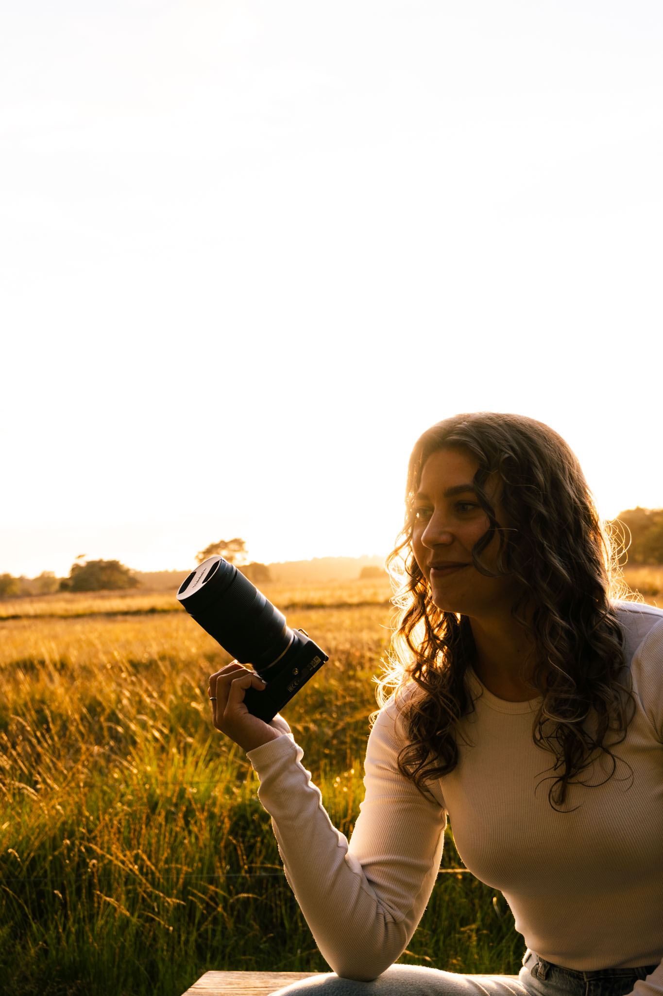 fotograaf kijkend naar de zijkant. ze houd haar camera om hoog. ze zit op een houten bankje met een mooie gouden ondergaande zon achter haar
